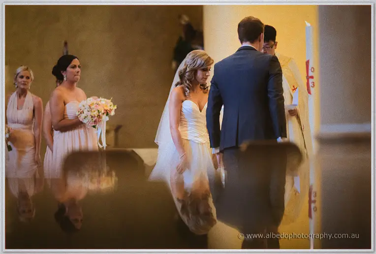 Cristina and Mitch stand with the priest at the altar as bridesmaids watch, soft reflections below