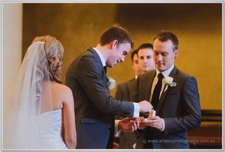 The best man passes the wedding rings to Mitch as groomsmen watch in the basilica's warm golden light
