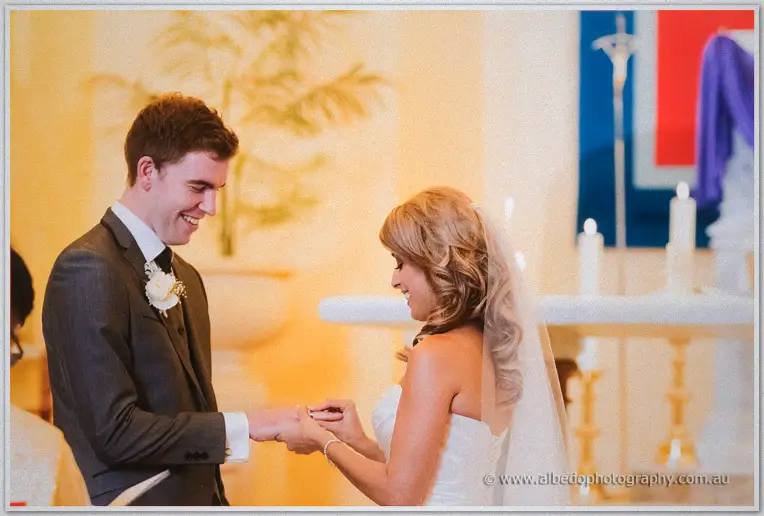 Mitch grins as Cristina slides the ring onto his finger, candlelight and greenery soft behind them