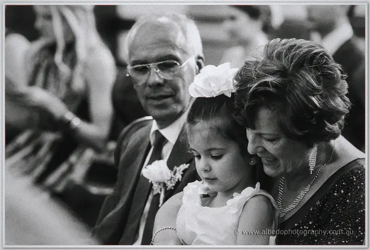 Black-and-white portrait of a flower girl between her grandparents, quiet and tender in the pews
