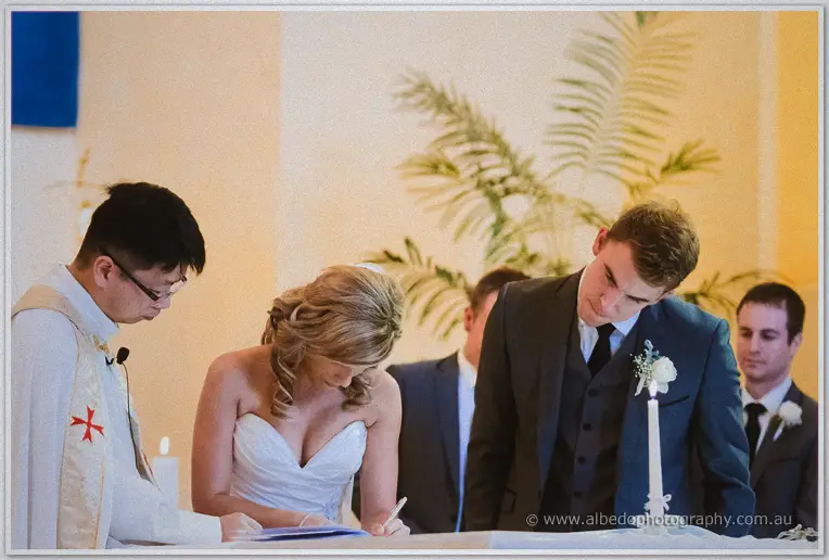 Cristina signs the marriage register at the altar with Mitch and the priest leaning close in candlelit warmth