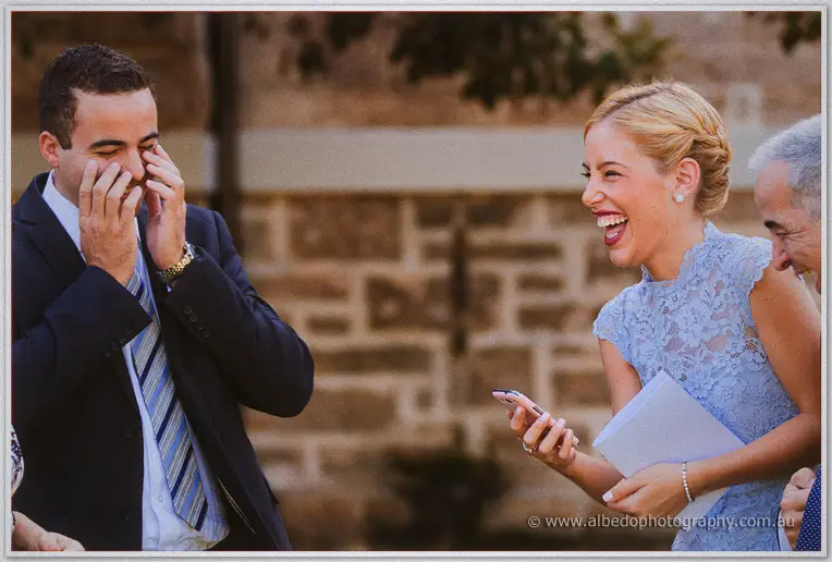 A groomsman covers his face laughing as a bridesmaid in powder-blue cackles outside the stone church