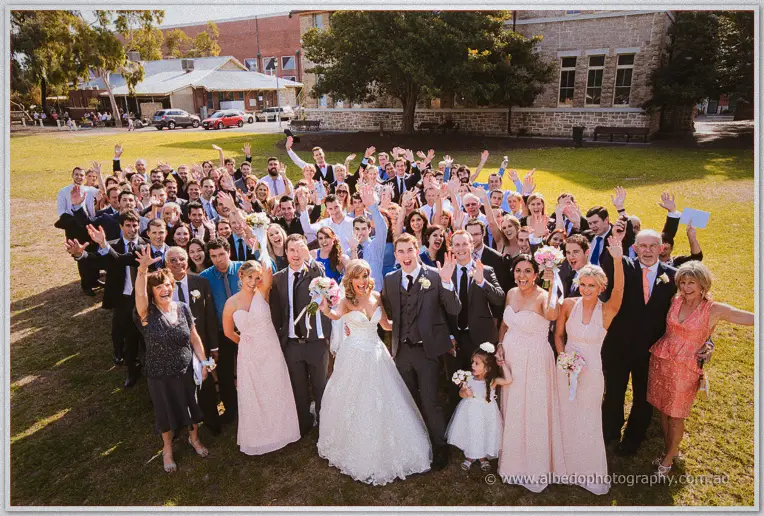 Crowd of wedding guests on a sunlit lawn waving above Cristina and Mitch's beaming family group
