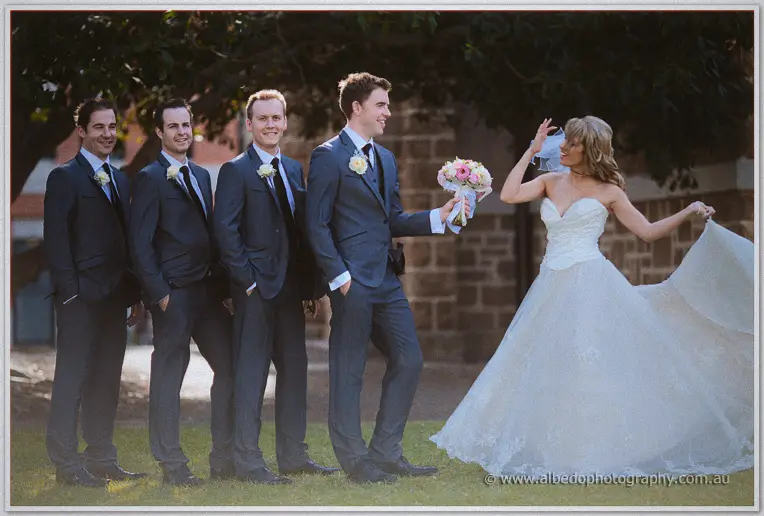 Mitch offers Cristina his bouquet as she whirls past the grinning groomsmen by the stone church