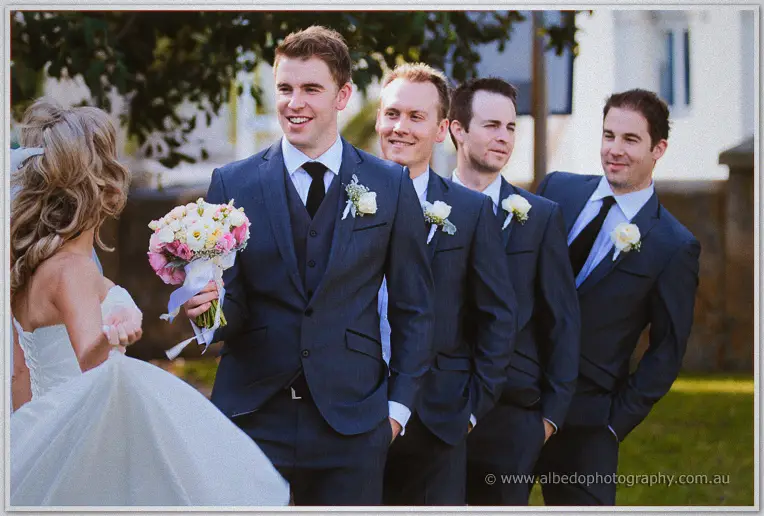 Groomsmen in navy suits line up laughing as Cristina bustles past with her bouquet outside St Patrick's