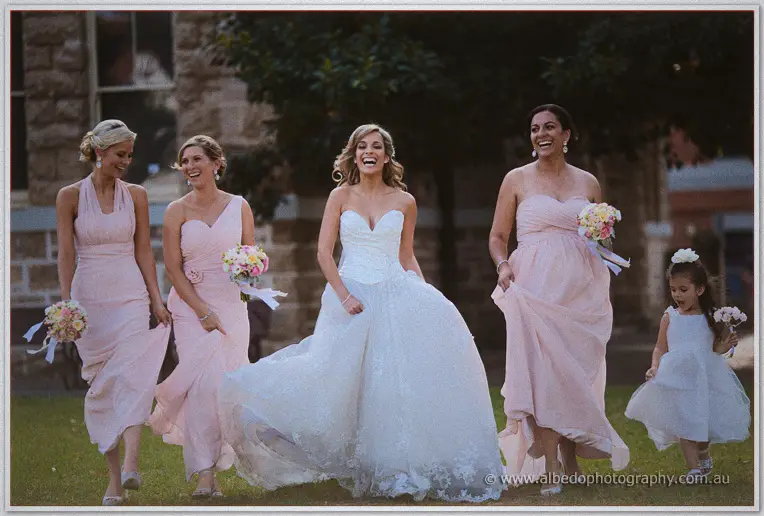 Cristina strides across the lawn lifting her gown, blush bridesmaids and flower girl sweeping behind