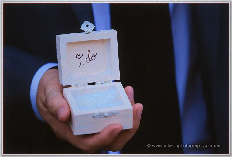A groom's hand opens a pale timber ring box inscribed with a heart and 'i do' in moody blue light