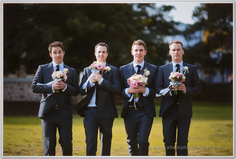 Four groomsmen in charcoal suits clutch pink posies to their chests beneath the trees, deadpan and amused