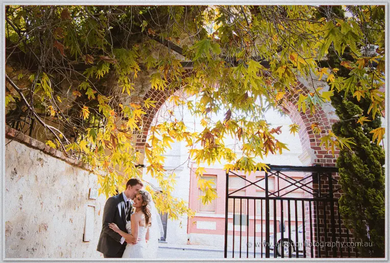 Cristina and Mitch embrace beneath a canopy of golden leaves by a brick archway
