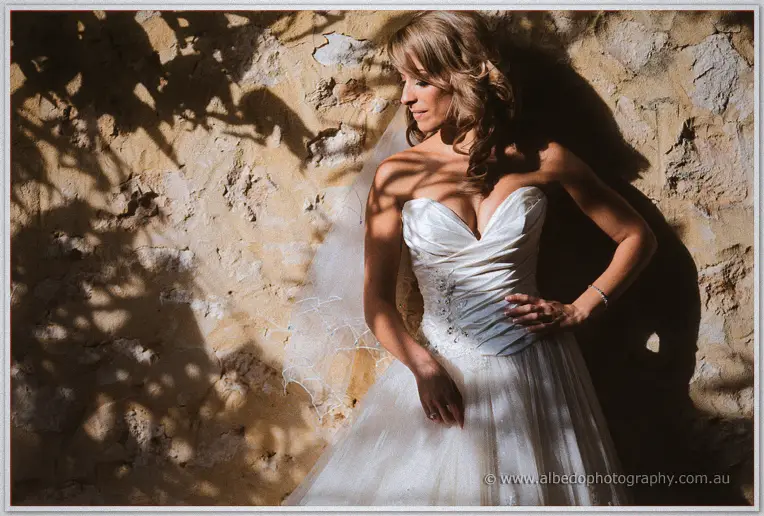 Cristina poses alone against a sun-dappled limestone wall, strapless gown and veil catching the light