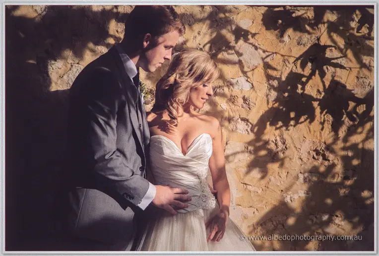 Mitch wraps Cristina in his arms before the golden stone wall, leaf shadows rippling across them