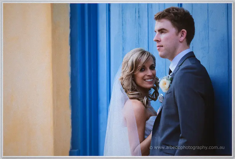 Cristina smiles past the camera beside Mitch beneath a weathered blue timber door