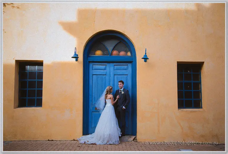 Wide shot of Cristina and Mitch before an arched blue doorway in a sunbaked ochre facade