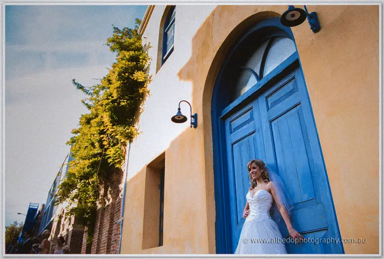 Cristina leans on the blue arched door under a bold sky as guests watch from the street below