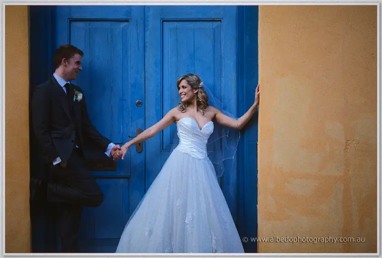 Cristina tugs Mitch's hand playfully before the vast blue doors, both laughing in the amber light