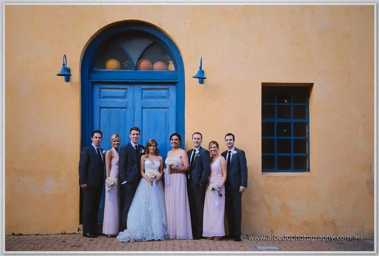 Full bridal party of eight poses before the blue arched door, blush gowns and soft evening sun