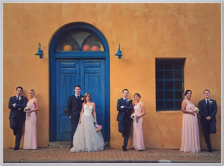 Bridal party pairs lean along the ochre wall either side of Cristina and Mitch at the blue door