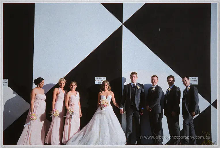 Bridal party lines up against a black-and-white geometric mural, Cristina and Mitch holding hands centre
