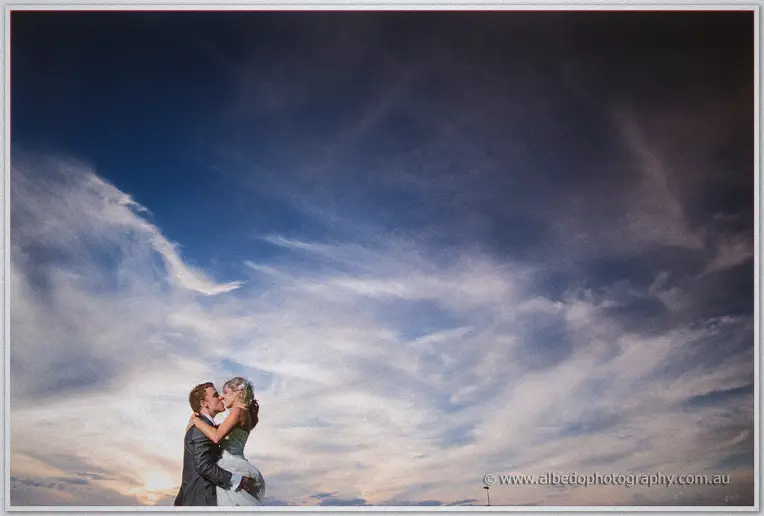 Cristina and Mitch kiss low in the frame beneath a vast swirling dusk sky after their Basilica ceremony
