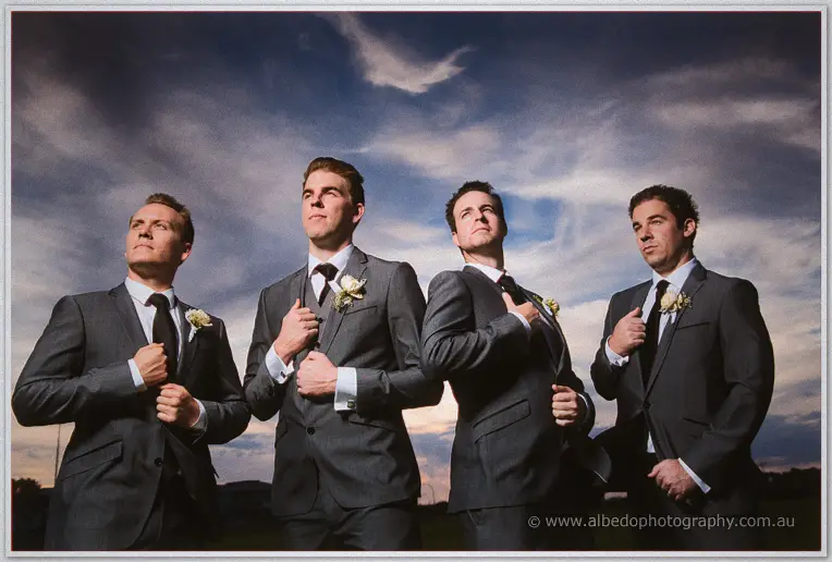 Four groomsmen in grey suits and white buttonholes straighten their jackets beneath a moody evening sky