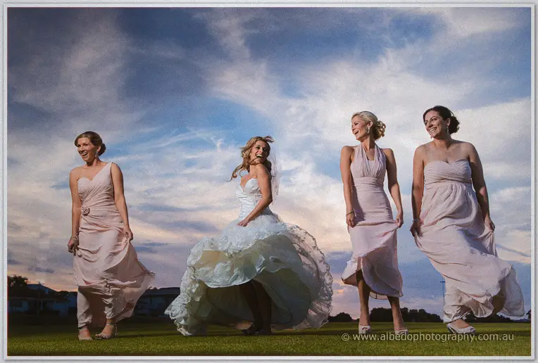 Cristina and three bridesmaids lift their skirts and laugh as wind whips them across the oval at sunset