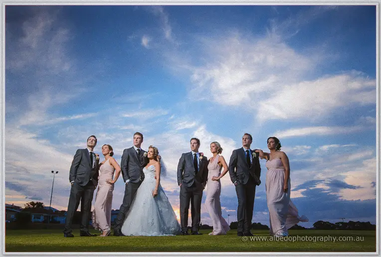 Bridal party in pink gowns and grey suits lines the grass beneath a deep blue cloudy twilight sky