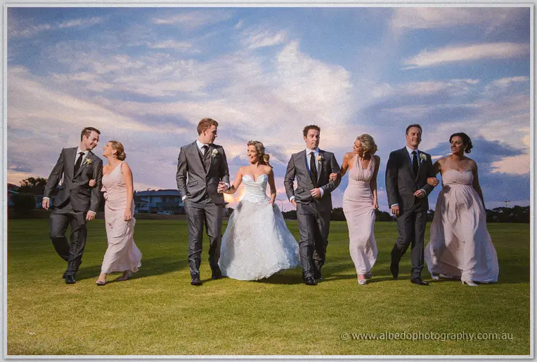 The full bridal party walks arm in arm across the green, Cristina lifting her dress mid-stride at dusk