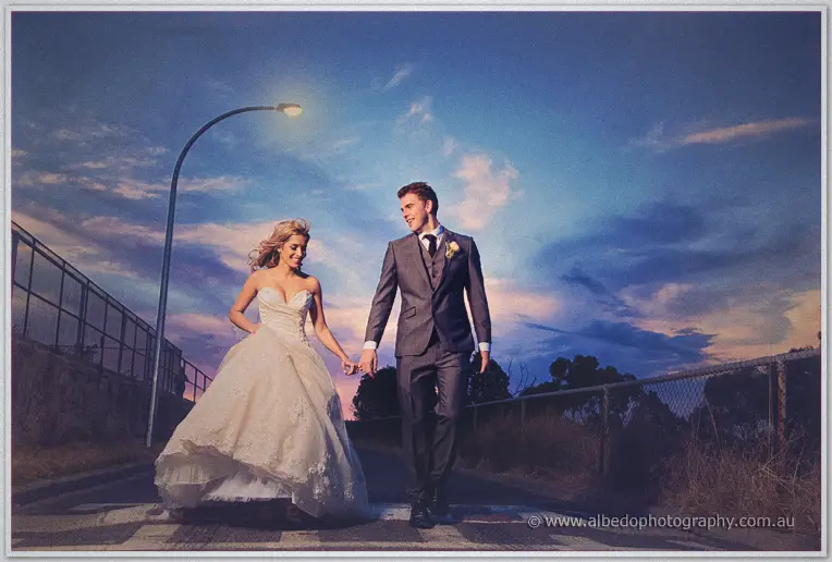 Cristina and Mitch cross a quiet suburban road hand in hand beneath a glowing streetlight at twilight