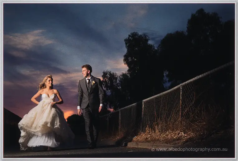 Cristina hauls her lace gown along a darkening path past a wire fence, Mitch smiling at her side