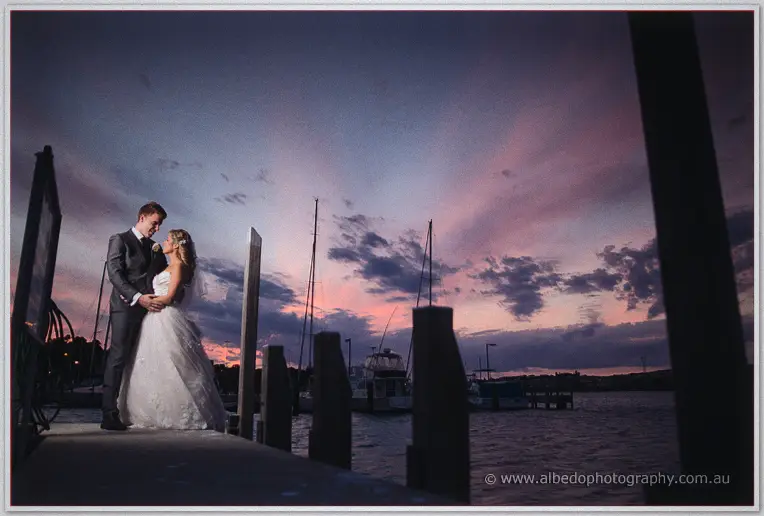 Cristina and Mitch embrace on a marina jetty framed by dark pylons against a pink and violet sunset