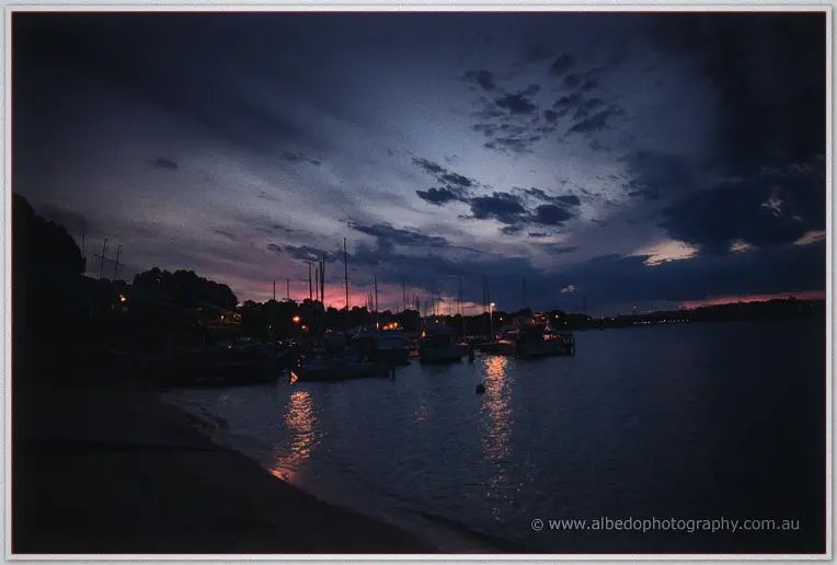 Night falls over the yacht club marina, moored boats silhouetted as harbour lights shimmer on the water
