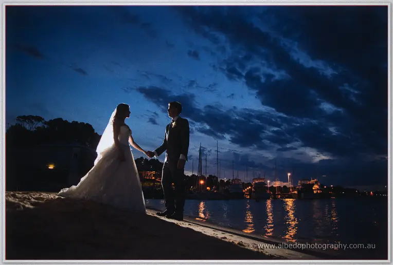 Bride and groom hold hands facing each other on the river beach as moored yachts glow behind them