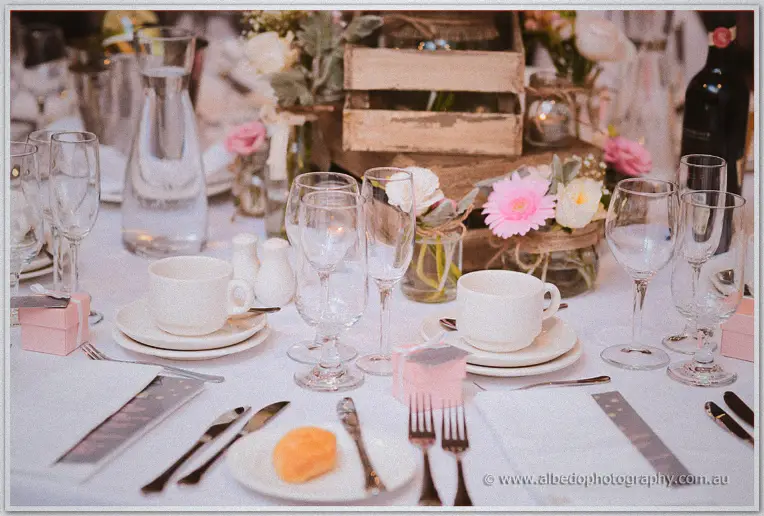 A crisp white table setting with teacups, wine glasses and pink favour boxes awaits guests at the club