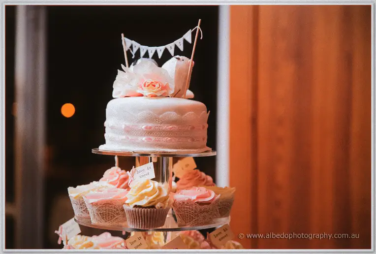 A lace-detailed cake topped with blooms and a LOVE bunting sits atop a stand of pastel cupcakes