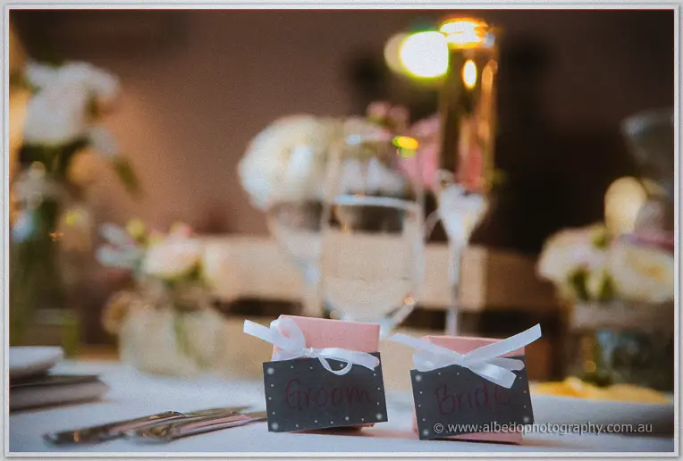 Handwritten bride and groom favour boxes tied with ribbon sit on the table in soft candlelit focus
