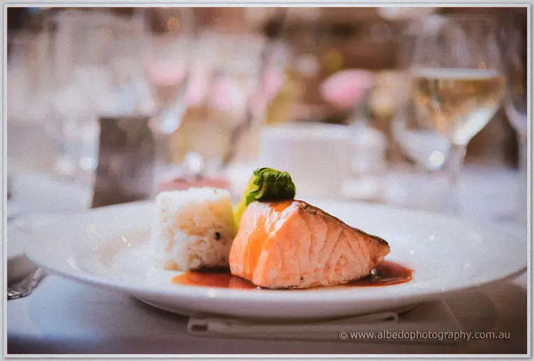 A plated salmon fillet with rice and greens gleams under soft light amid wine glasses and blurred blooms