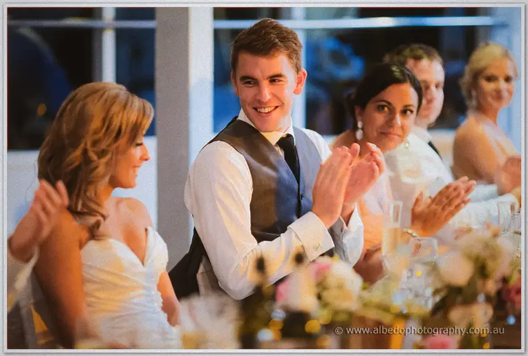 Groomsmen in grey vests clap and laugh beside Cristina at the candlelit bridal table during speeches