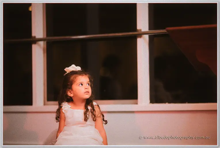 A young flower girl in a white dress with a rose headband gazes wide-eyed across the reception room