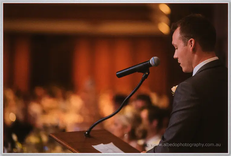 A groomsman in a dark suit delivers his speech into a microphone before glowing, blurred guests