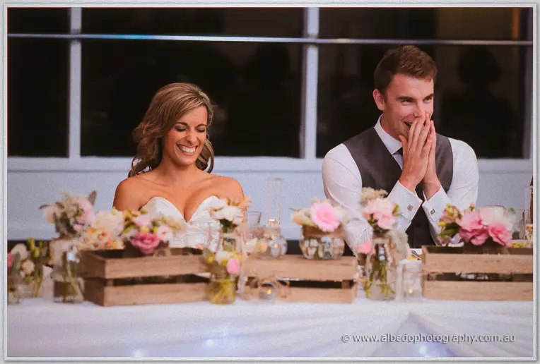 Cristina giggles while Mitch covers his mouth laughing behind crate arrangements at the bridal table