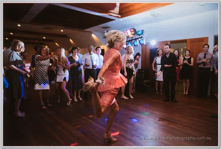 A guest in a coral dress kicks up her heel dancing while friends cheer around the lit dance floor