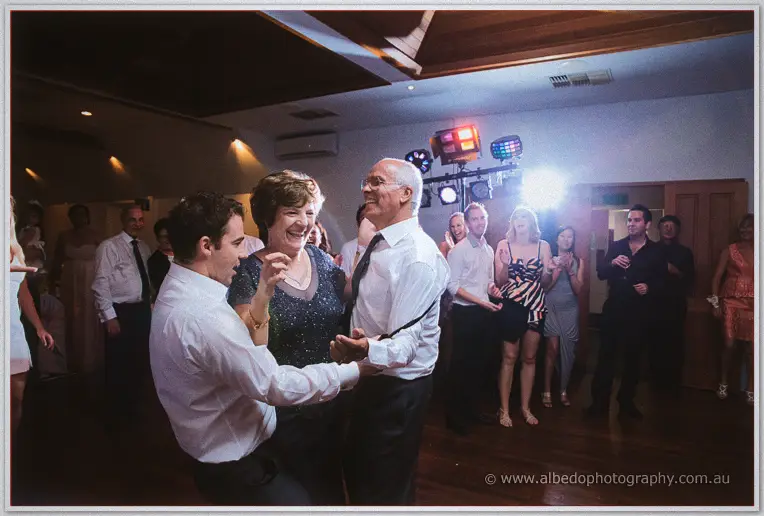 Two guests swing a laughing older woman between them on the dance floor under party lights