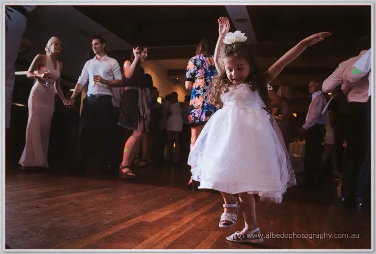 A little girl in a white frock with a flower clip twirls arms raised on the crowded dance floor