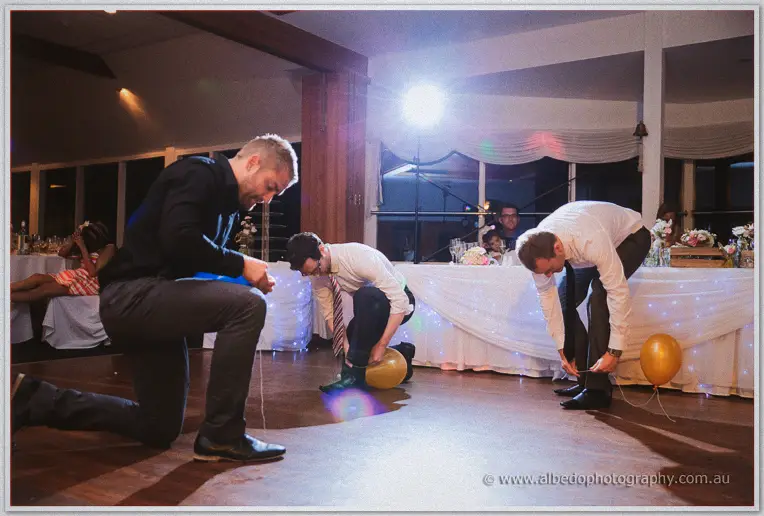 Three men kneel on the dance floor tying coloured balloons to their ankles for a party game