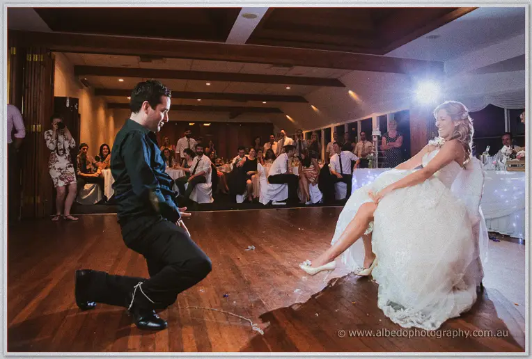 A groomsman kneels before seated Cristina, reaching for the garter as guests watch the tradition
