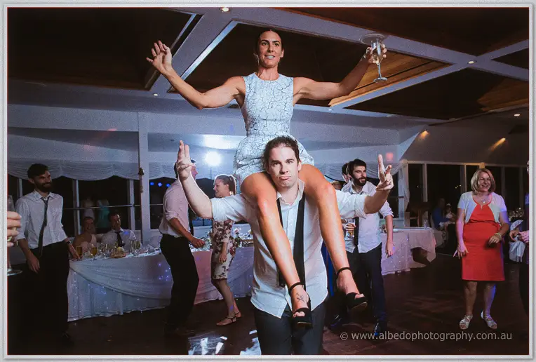 Groom carries a champagne-holding woman in white lace on his shoulders at the yacht club reception