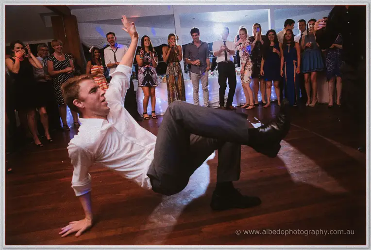 Groomsman in white shirt and grey trousers breakdances mid-move for laughing guests at the reception