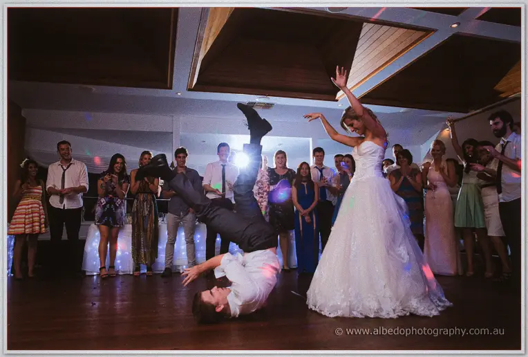 Bride in strapless lace gown dances beside a cartwheeling groomsman under disco lights at East Fremantle