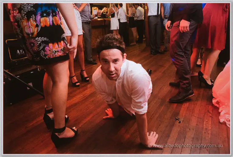 Guest in white shirt crouches playfully on the timber dance floor amid dancing legs and warm bar light