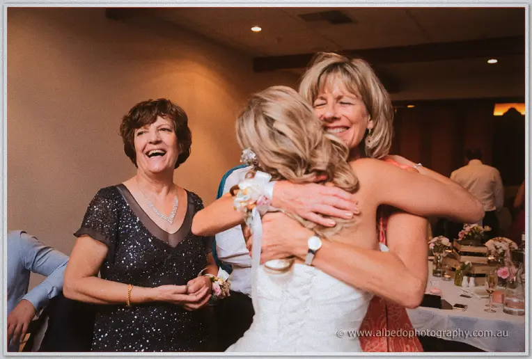 Bride hugs a beaming older woman in coral while another guest laughs beside them at the reception